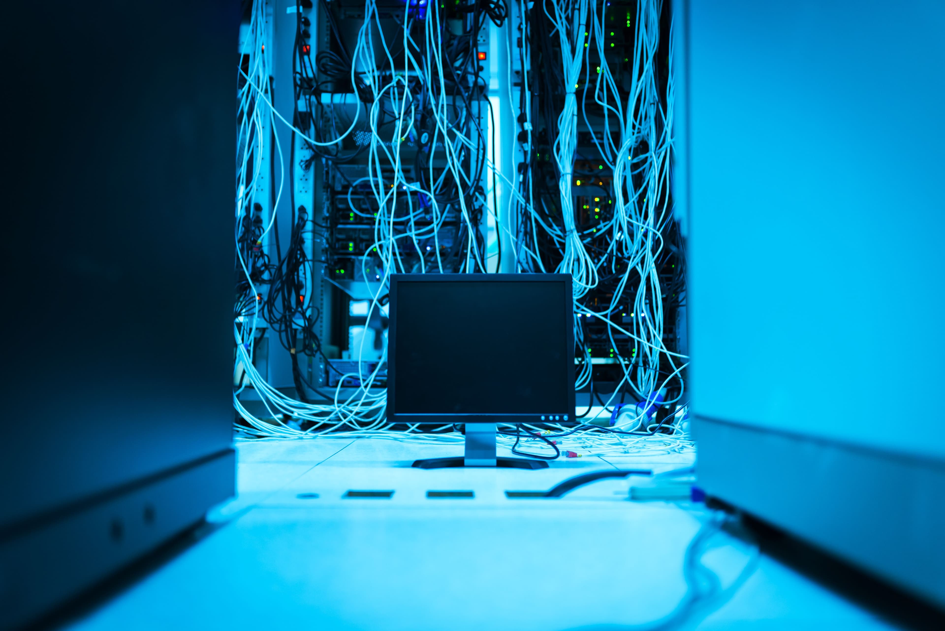 Tangled network cabling draped over server racks lit in blue, with a single monitor in the foreground